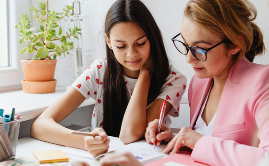 A young woman with long dark hair sits beside a woman with glasses and a pink jacket at a table. They both hold pens, and are working on schoolwork