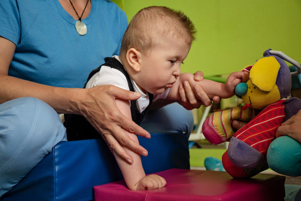 A young baby in dungarees is playing with toys, being supported by their carer