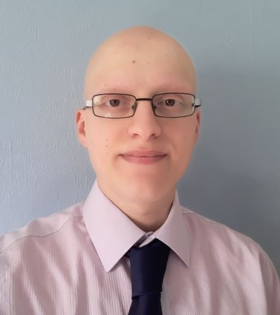Ali wears glasses and a pink shirt with a blue tie. He stands against a gray background and smiles softly.