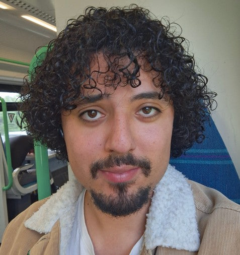 Anthony, a man with curly hair and a beard wearing a beige jacket with white collar sitting on a bus.