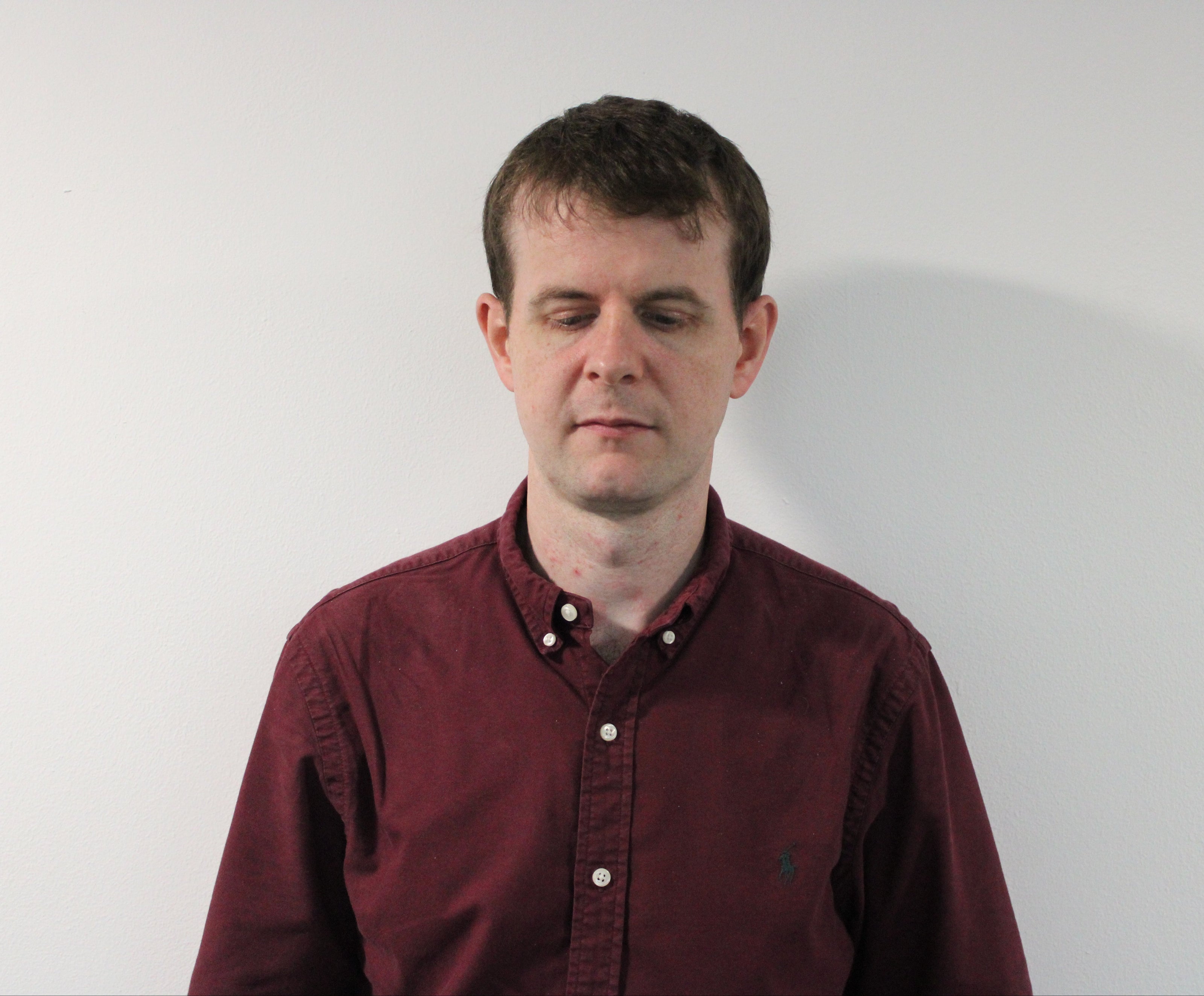 Callum, a man wearing a maroon button-up shirt standing against a plain white wall.