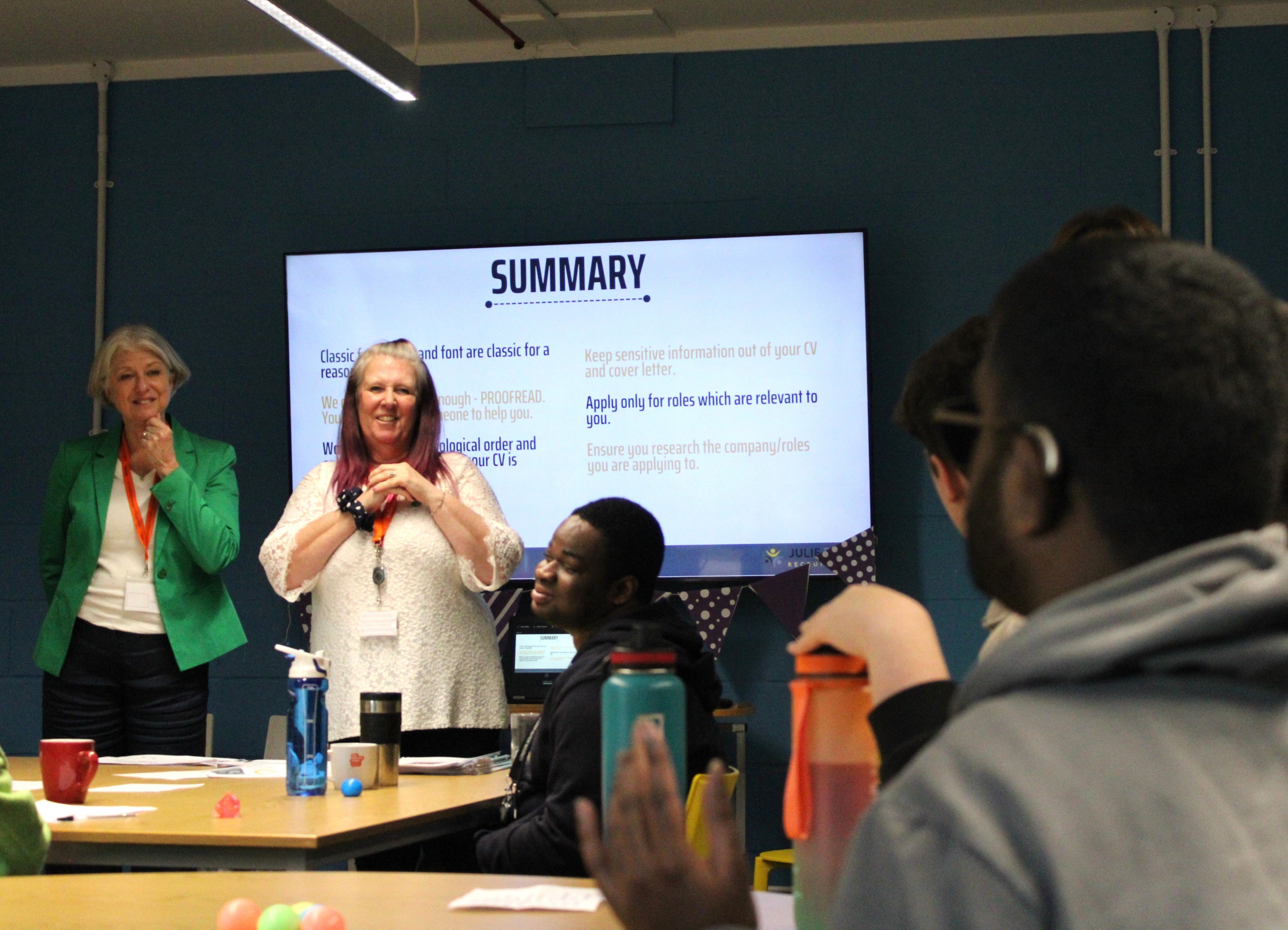 Two presenters stand in front of a screen talking through a summary presentation slide to a group of students in a classroom.