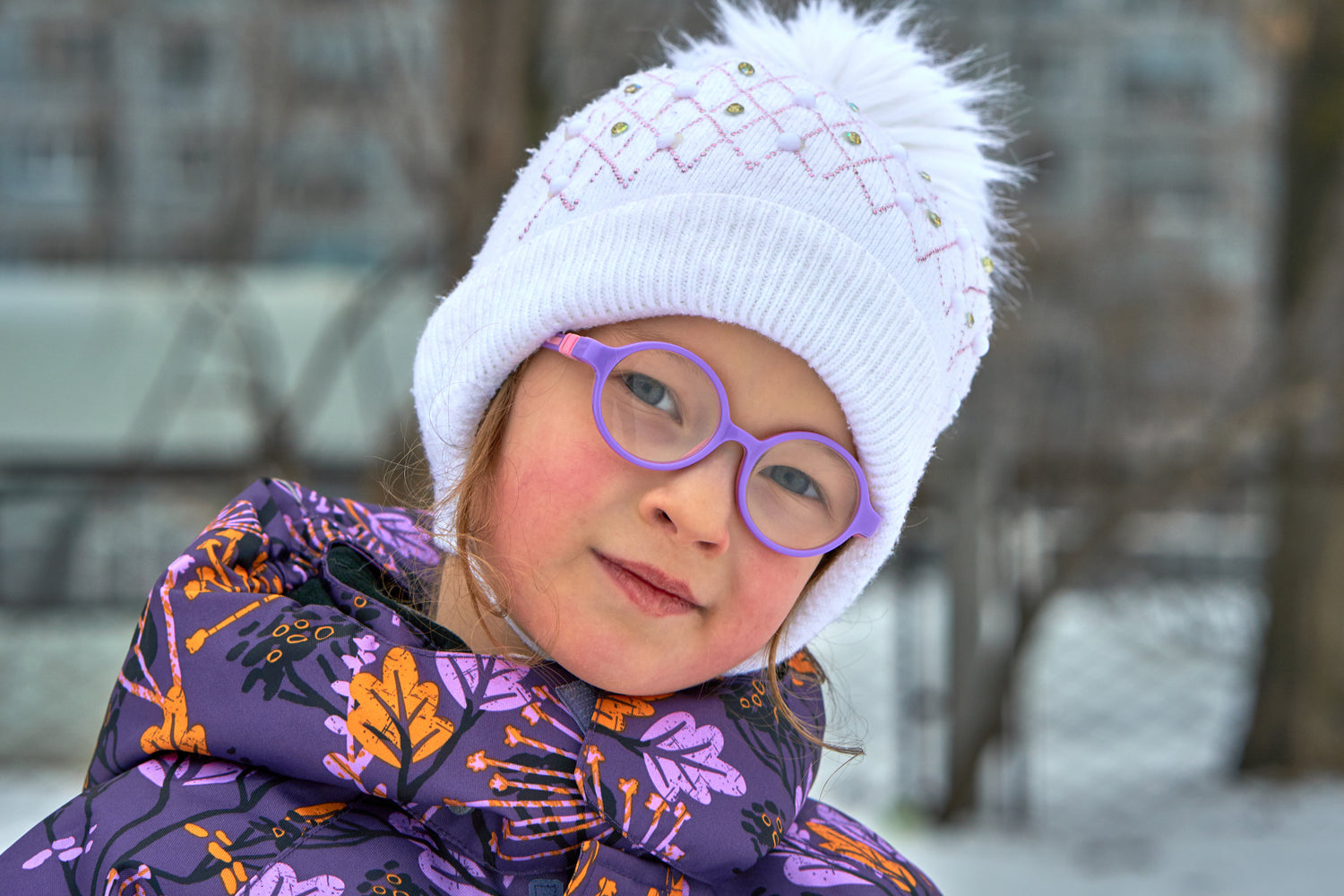 A child wearing purple glasses, a white knitted beanie and a colorful patterned jacket, standing outdoors in winter, smiling softly.