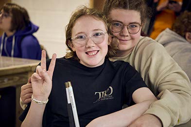 two smiling girls with glasses smiling at the camera; one has a cane and is making a peace sign
