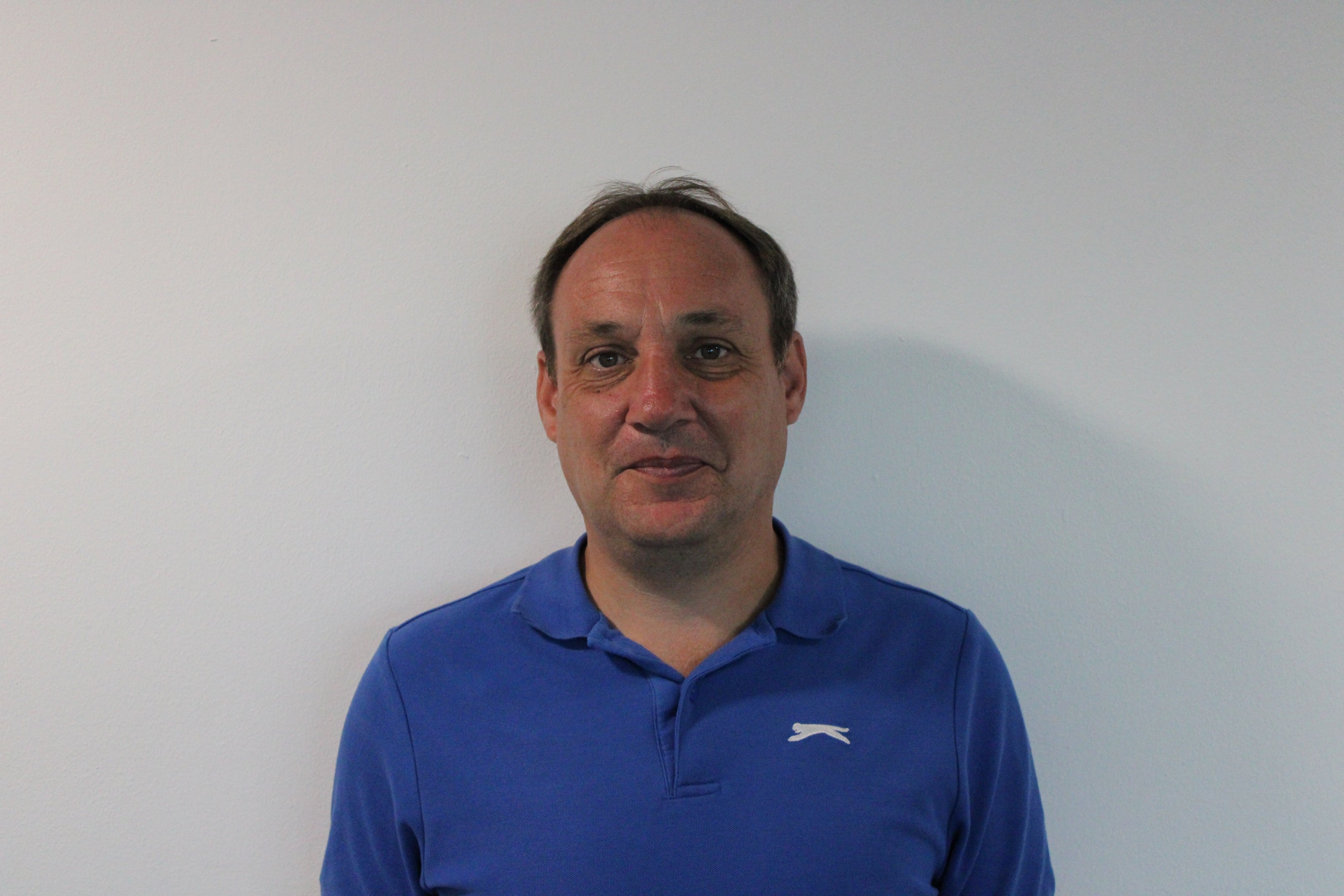 Alex, a man wearing a blue polo shirt with a logo against a plain background