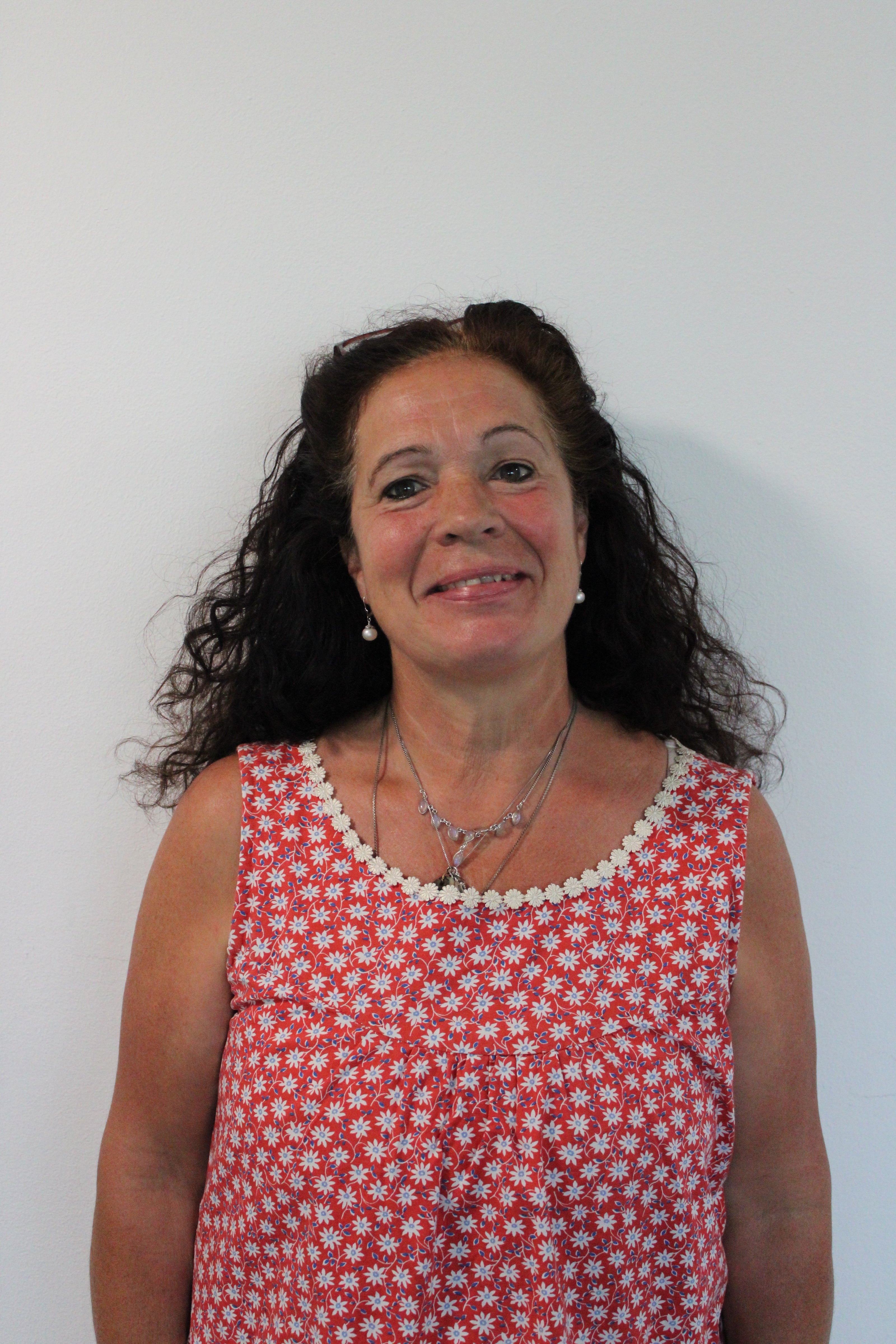 Alison, a woman with long curly dark hair wearing a sleeveless red dress with white daisy patterns standing against a plain white background.