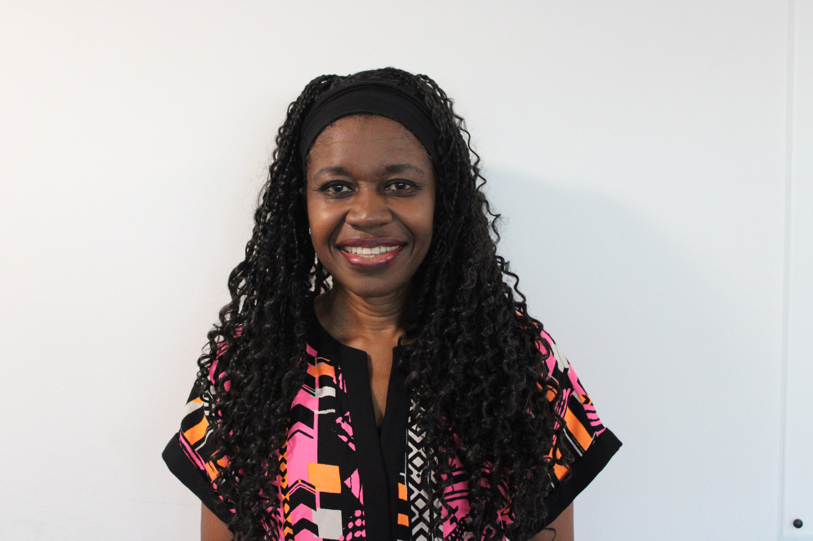 Ify, a woman with big smile and long dark hair wearing a colorful top against a white background.