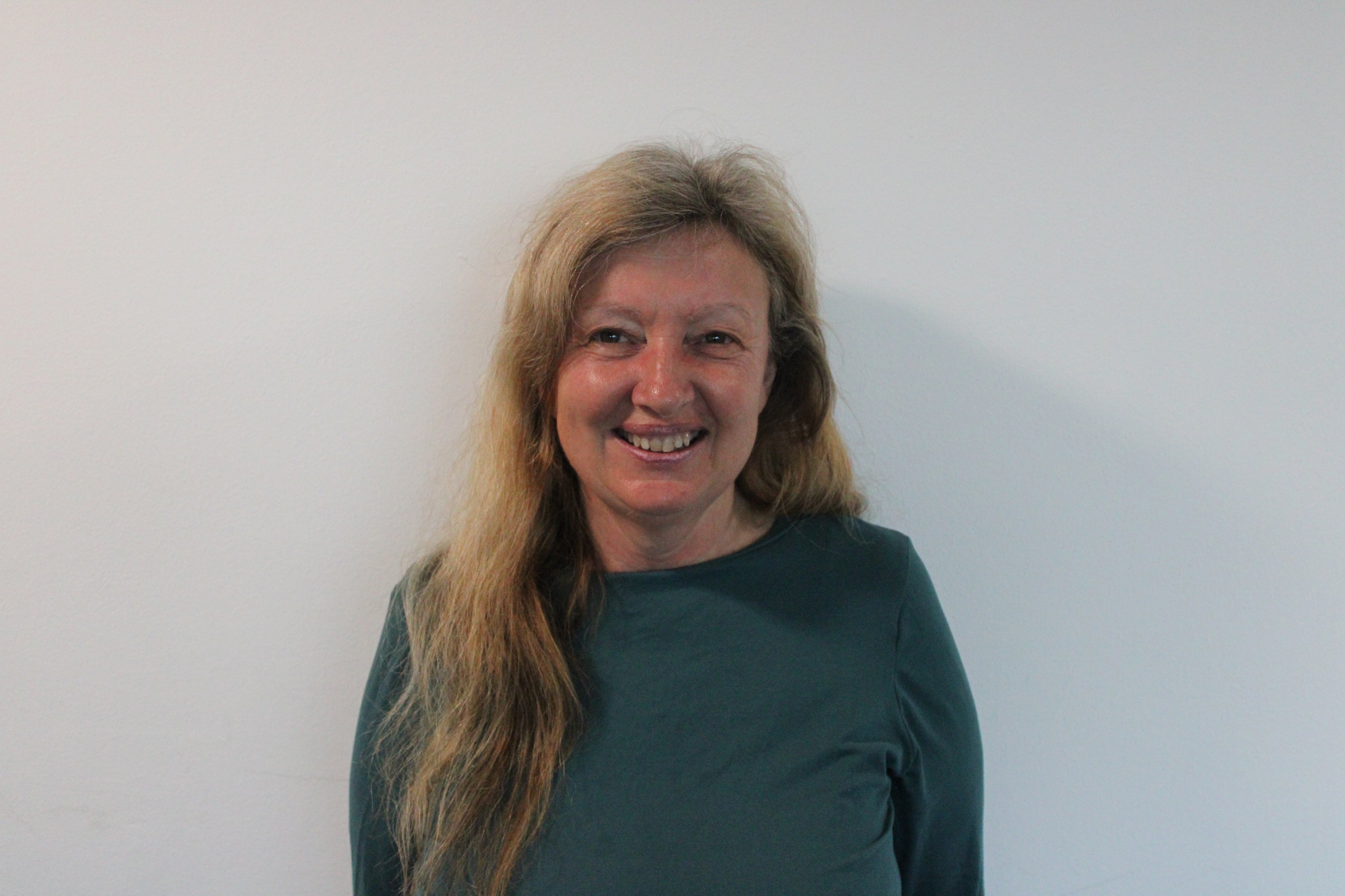 Janine, a woman with long blonde hair, wearing a dark green, long-sleeved top smiling standing against a white wall