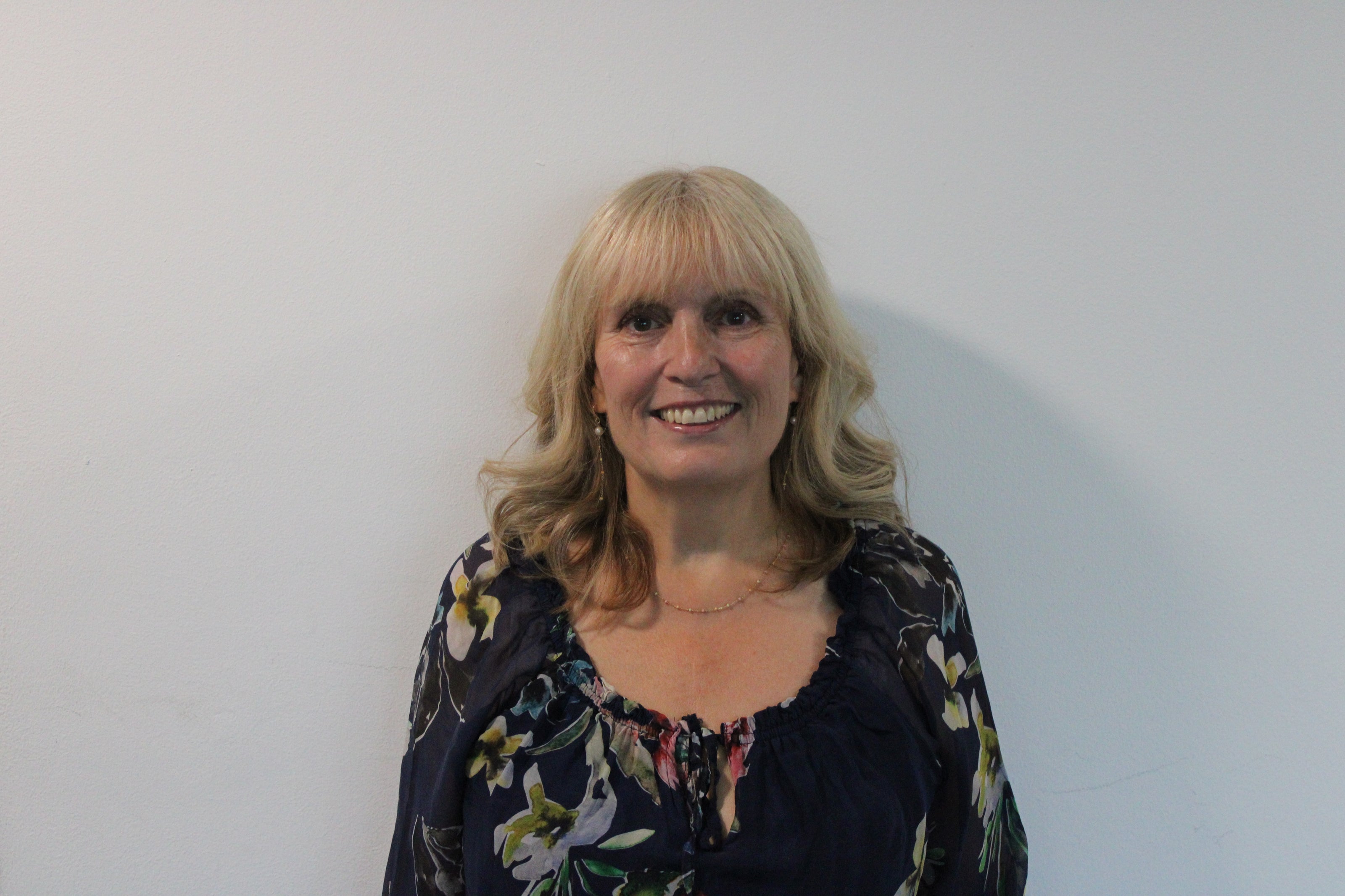 Lynne, a woman with blonde hair wearing a floral long-sleeved top smiling against a plain white background