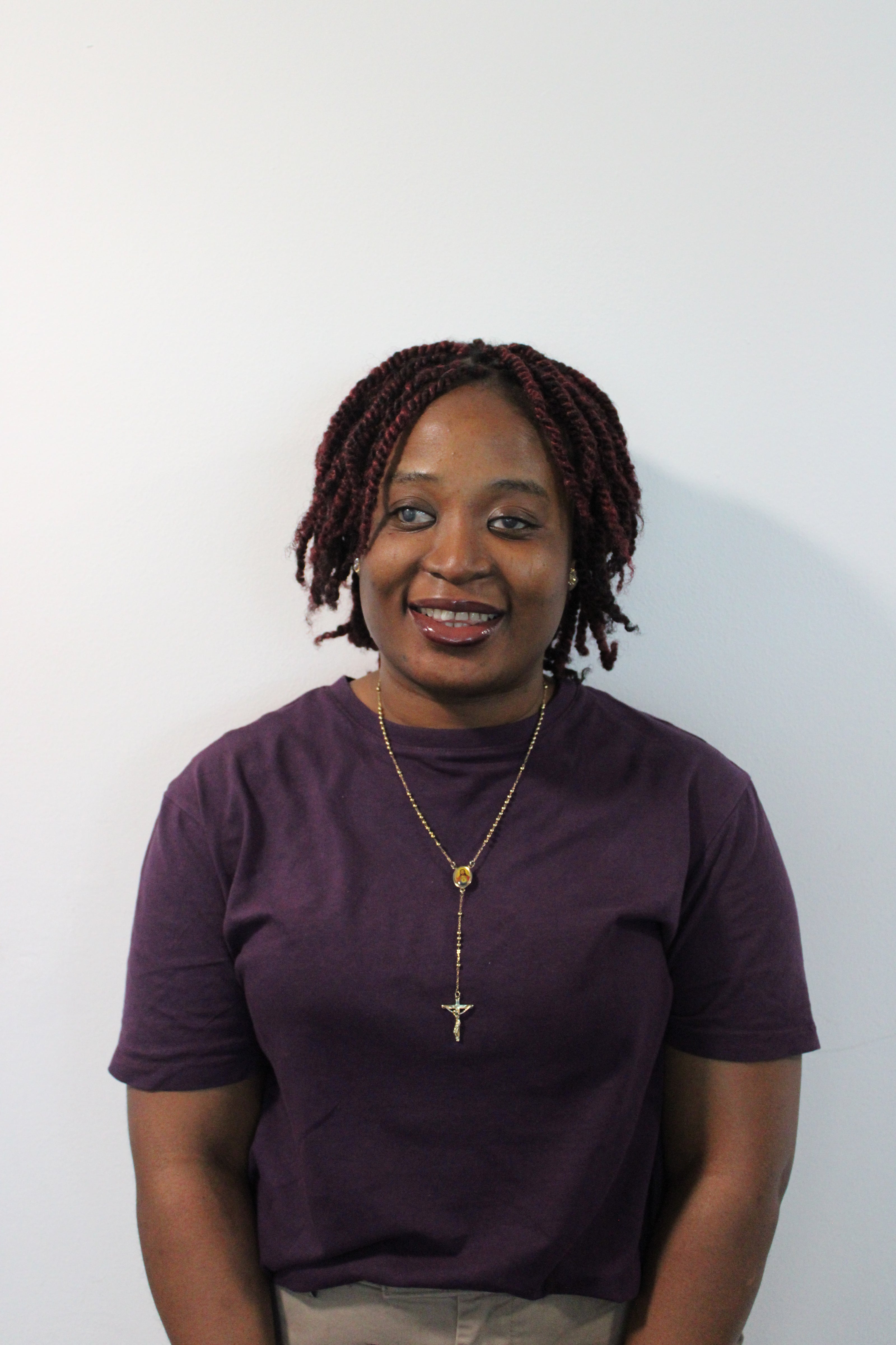 Monsurat, a women smiling, wearing a purple top, and a long gold necklace standing against a white wall