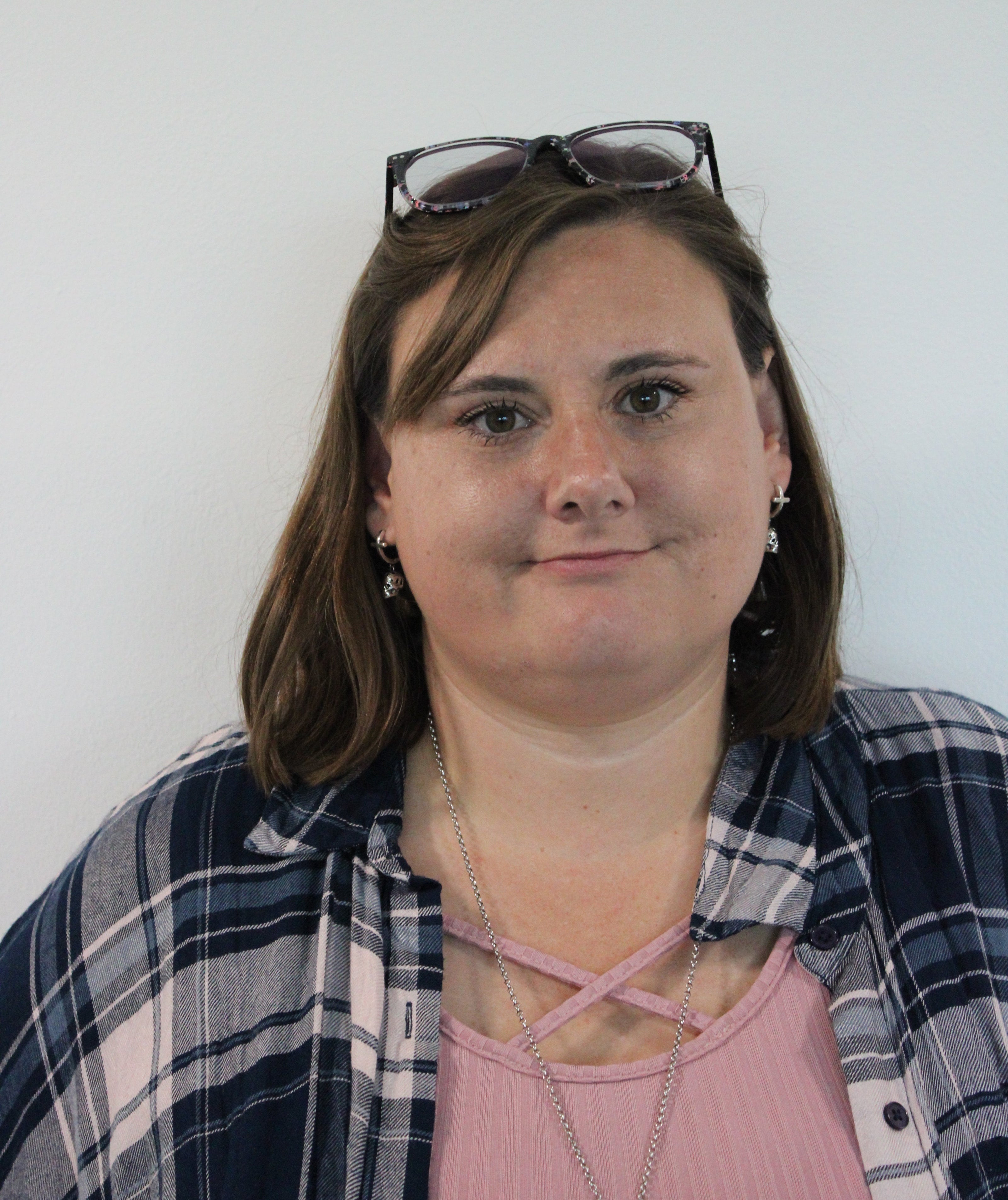 Sarah, a woman wearing a pink top and plaid shirt standing against a white background