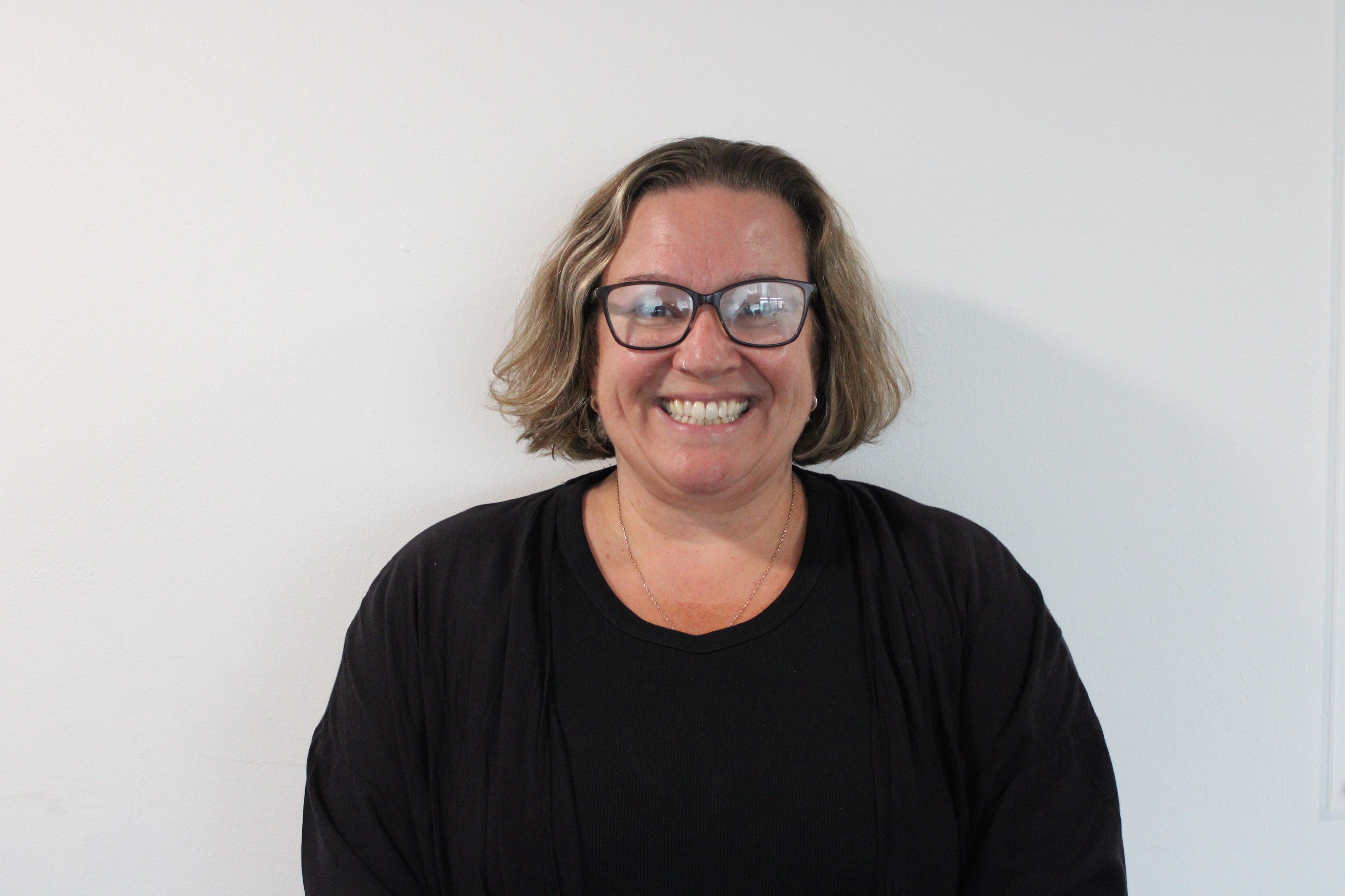 Toni, a woman smiling brightly, wearing glasses and a black jumper against a white background