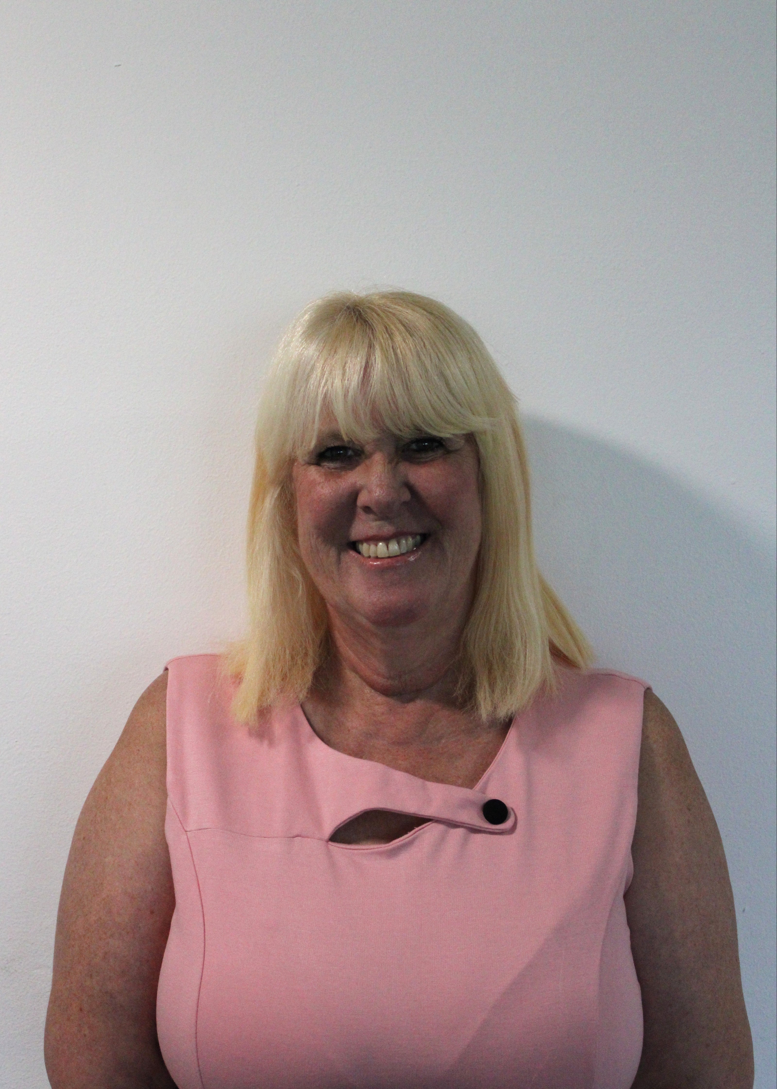 Tricia, a woman with blonde hair wearing a pink top against a white background
