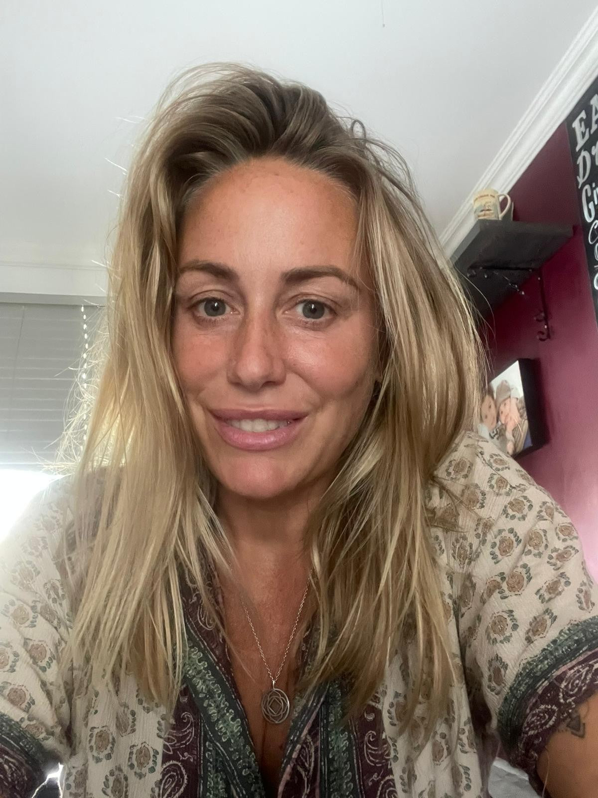 Emmeline, a woman with blonde hair wearing a printed top and silver necklace. She is in front of a window with white blinds and a burgundy wall with shelves and pictures on it.
