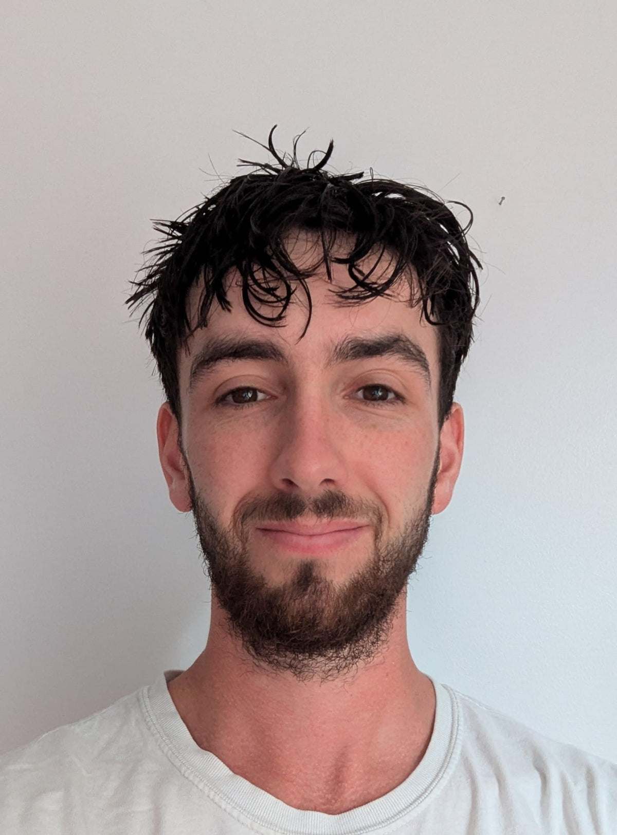 Close-up portrait of a young man with dark hair and a short beard smiling gently against a plain white background wearing a white t-shirt. 