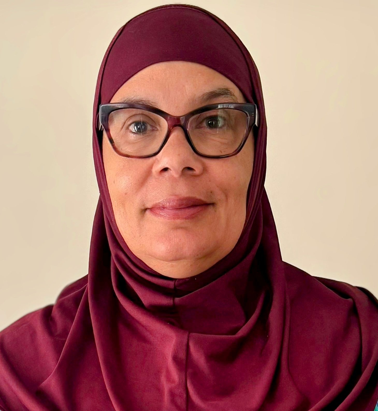 Woman wearing a maroon hijab and glasses against a beige background