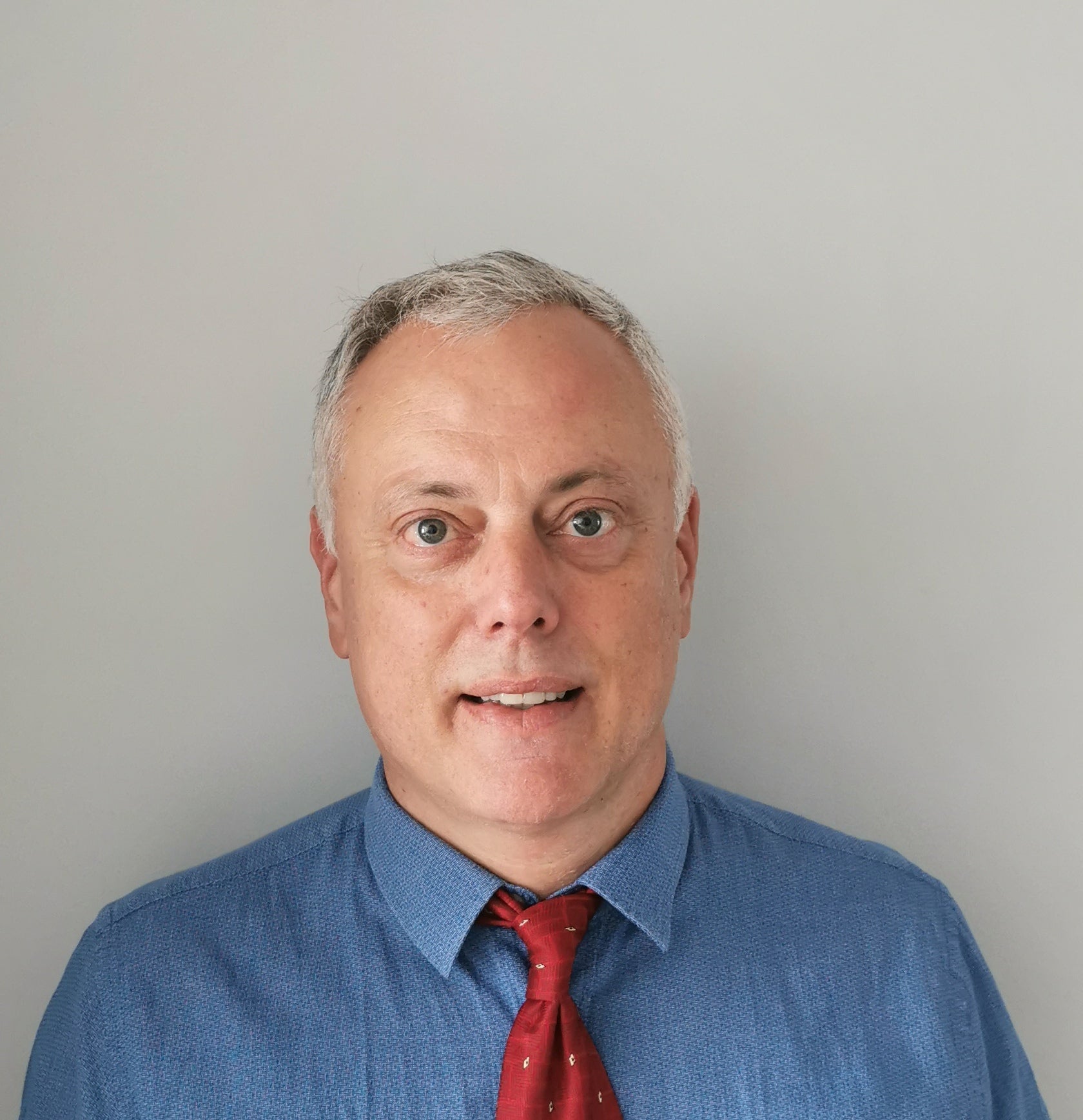 Man with grey hair wearing a blue shirt and red tie against a gray background