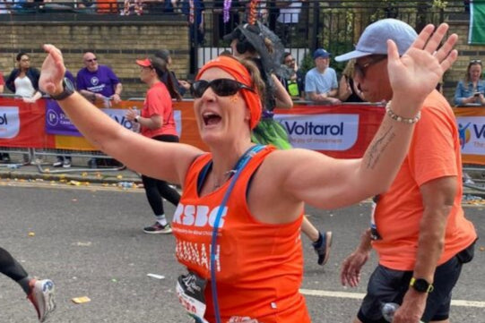 Hannah, RSBC runner in an orange vest celebrating in a marathon with spectators and flags in the background.