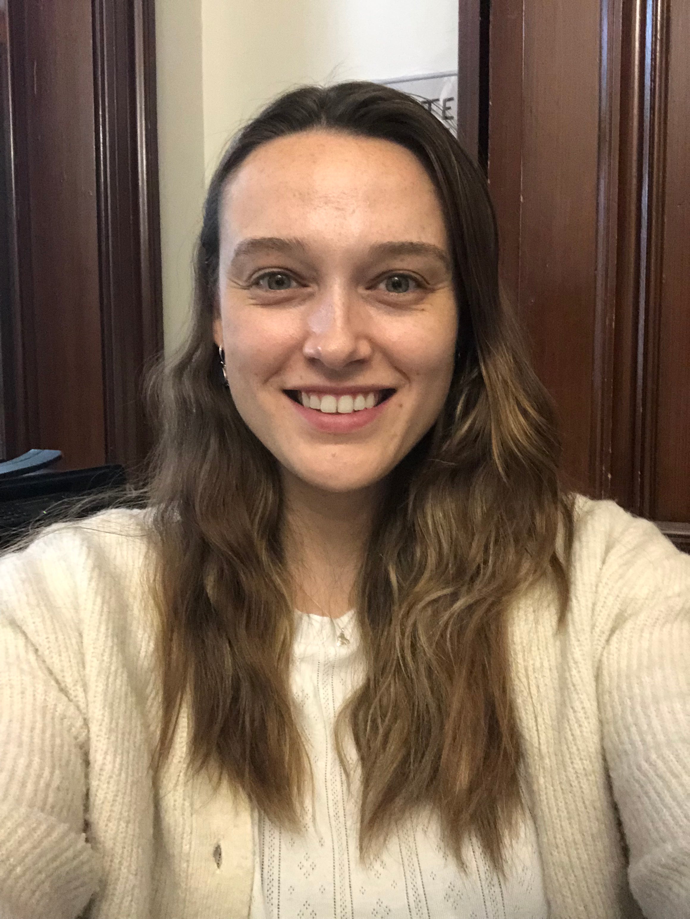 Harriet, a woman with long wavy brown hair wearing a white knit sweater, standing indoors against a wooden paneled wall.
