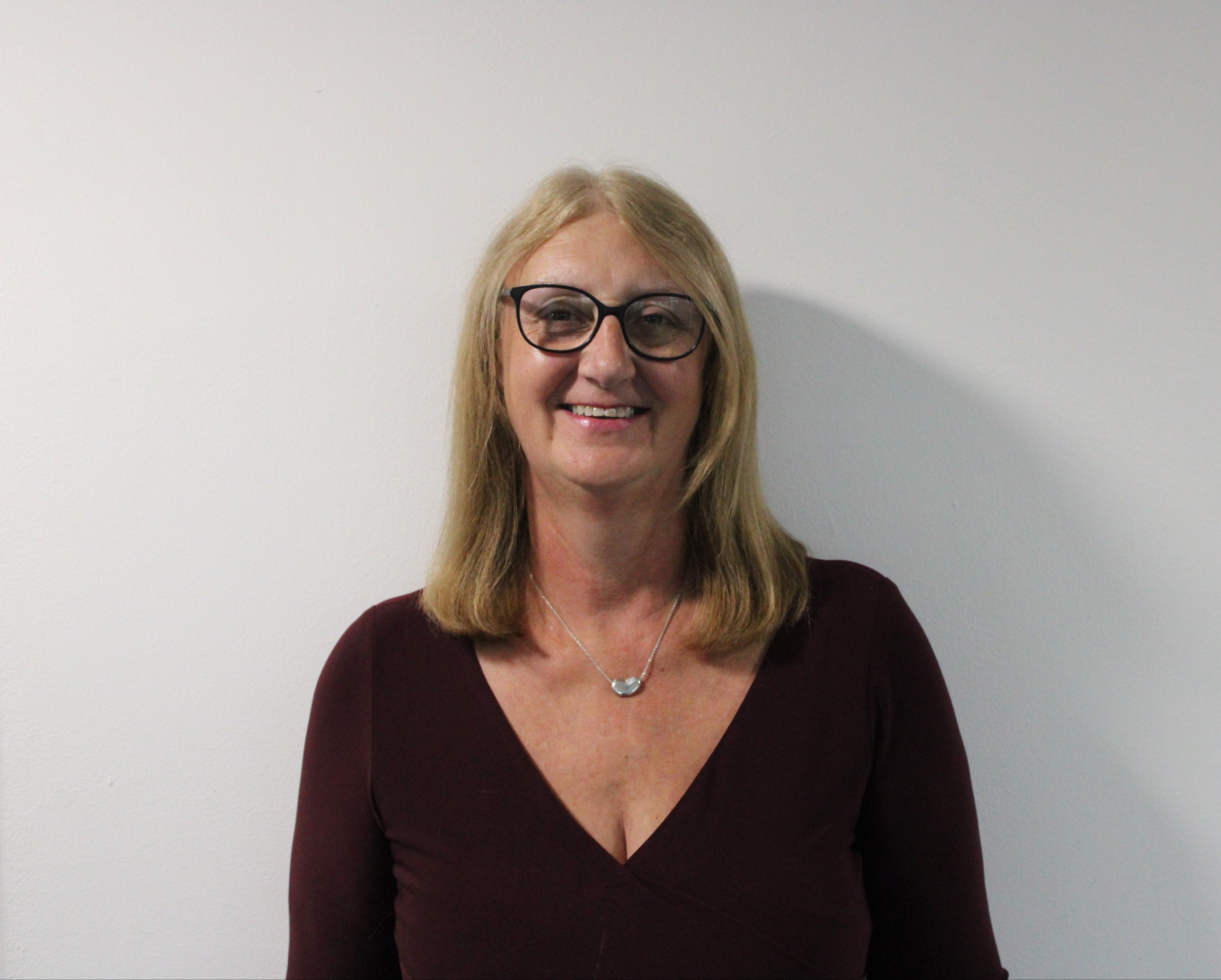 Janet, a woman with shoulder-length blonde hair wearing a dark burgundy V-neck top and a silver necklace against a plain white background.
