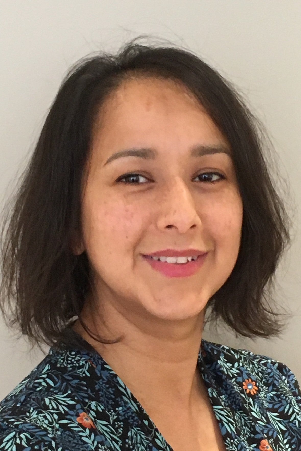 Smiling woman with shoulder-length dark hair wearing a floral patterned top against a plain background.
