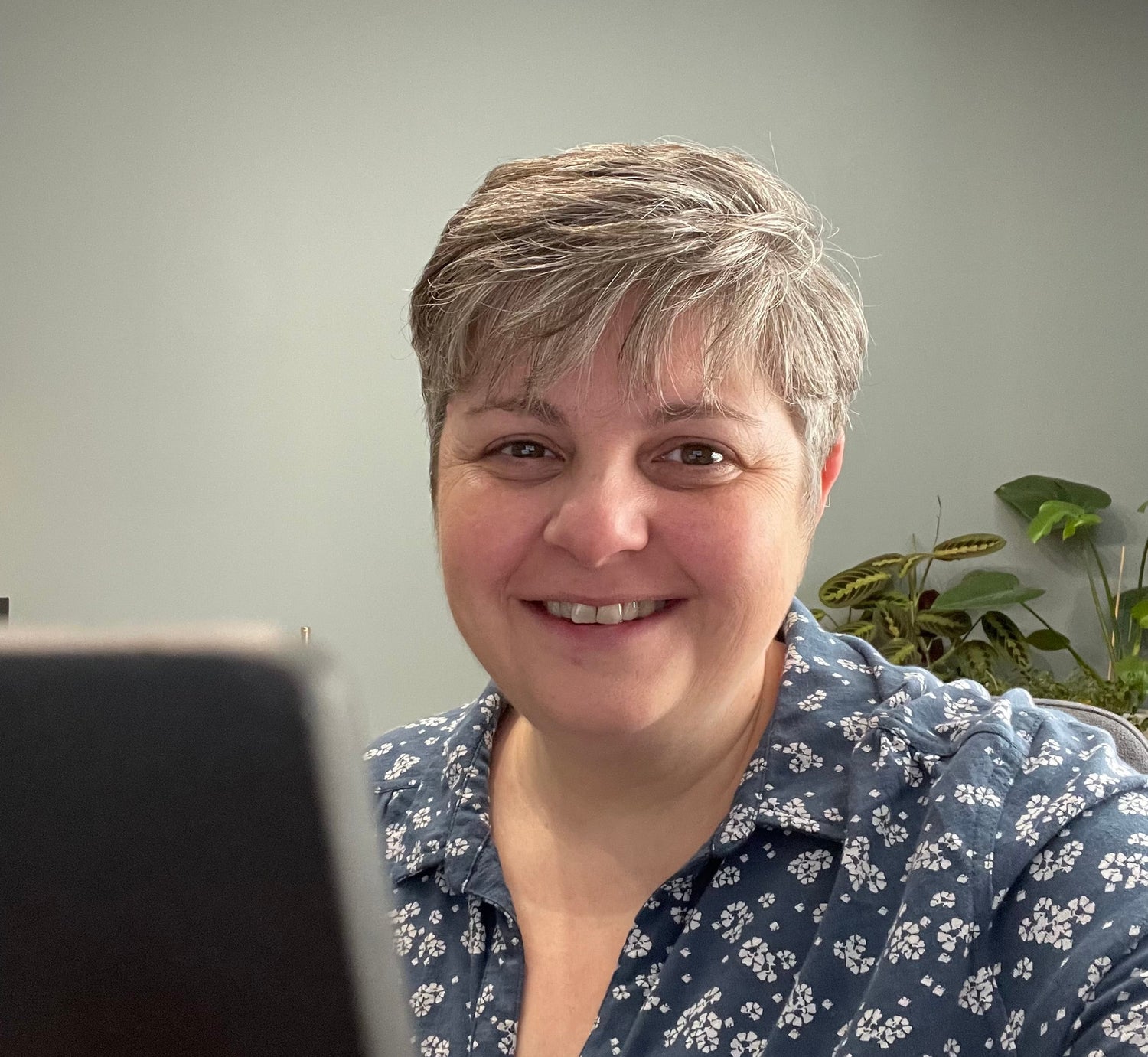 Jeanette, an RSBC Family Practitioner sitting in an office room smiling and facing the camera, wearing a greyish blue long-sleeved top covered in white flowers. A laptop is in front of her and a green plant behind. 