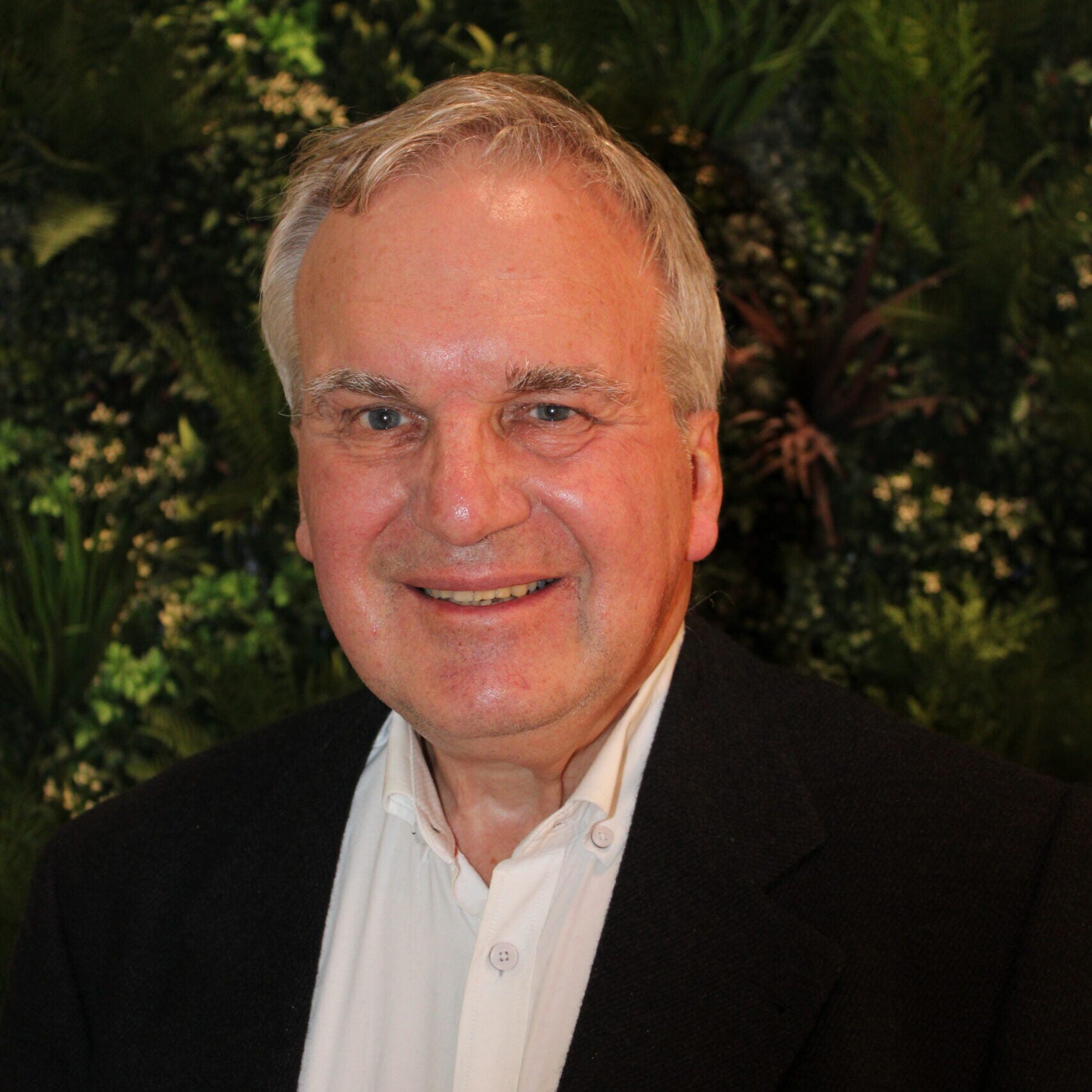 A smiling, clean shaven older man with white hair and a black suit