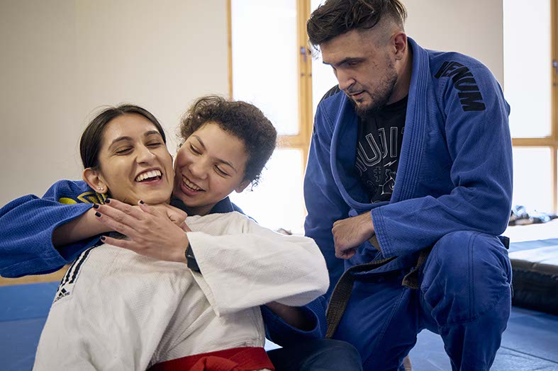 Two people practicing Judo on a blue mat with a teacher assisting.