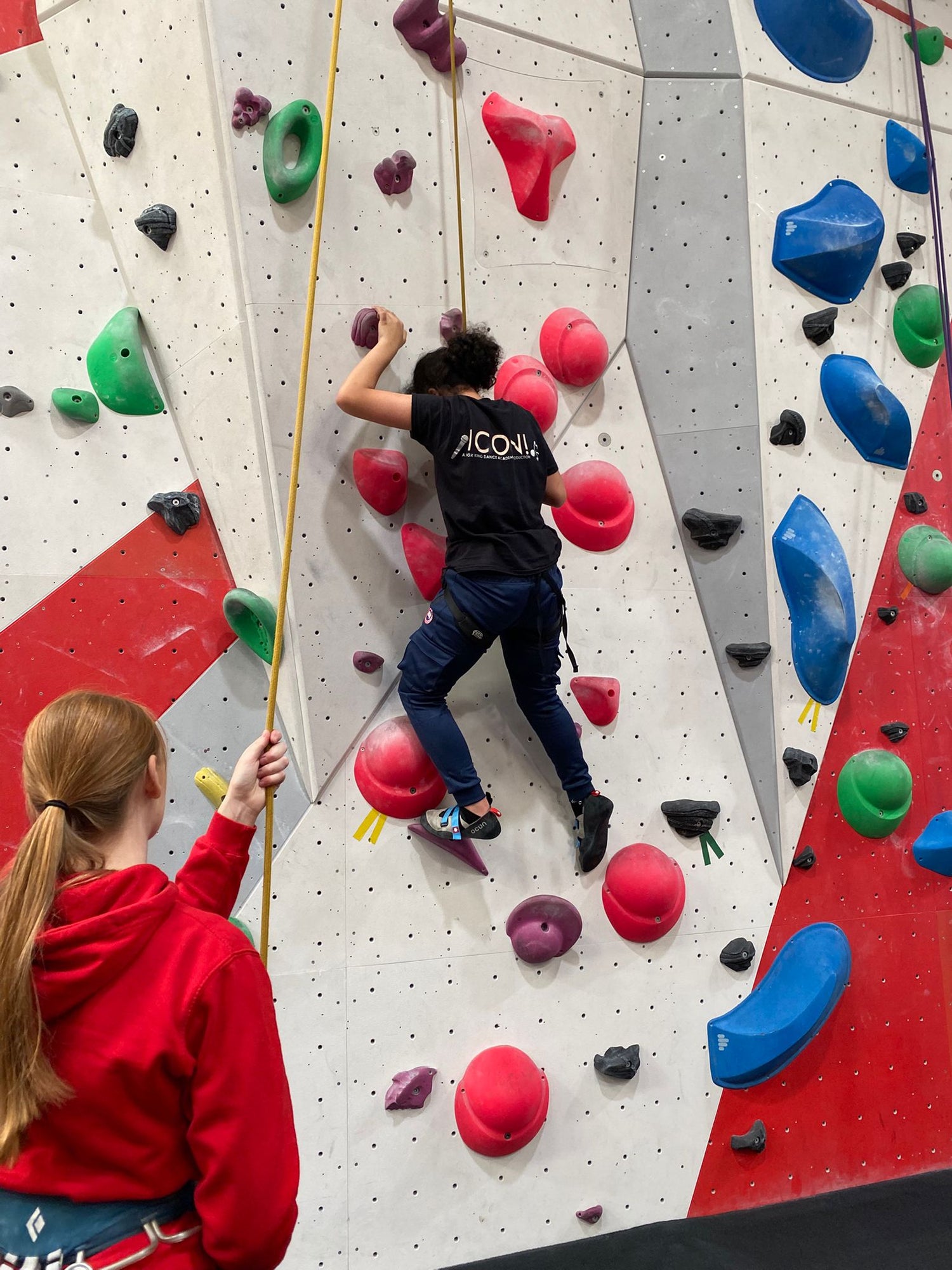 A climber ascends a colorful climbing wall while a belayer stands below, holding the rope for safety.