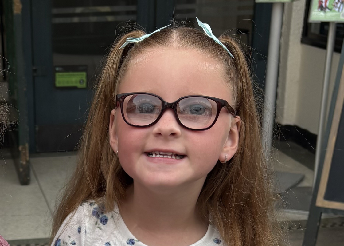 Maya, a young girl with blonde hair in pigtails wearing glasses. She's smiling sweetly at the camera.