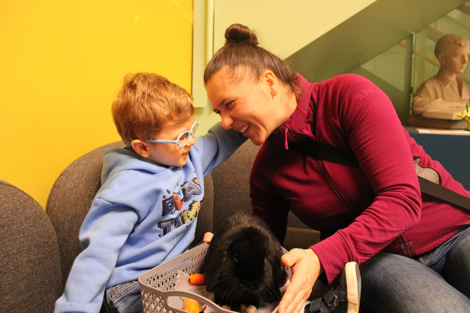 Mother and child sitting closely on chairs, gently petting a black rabbit in a basket filled with vegetables indoors.