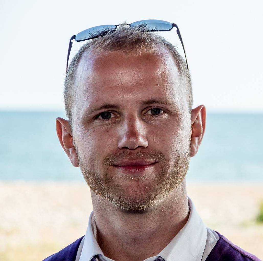 Man with a soft smile and sunglasses on head against a beach background.