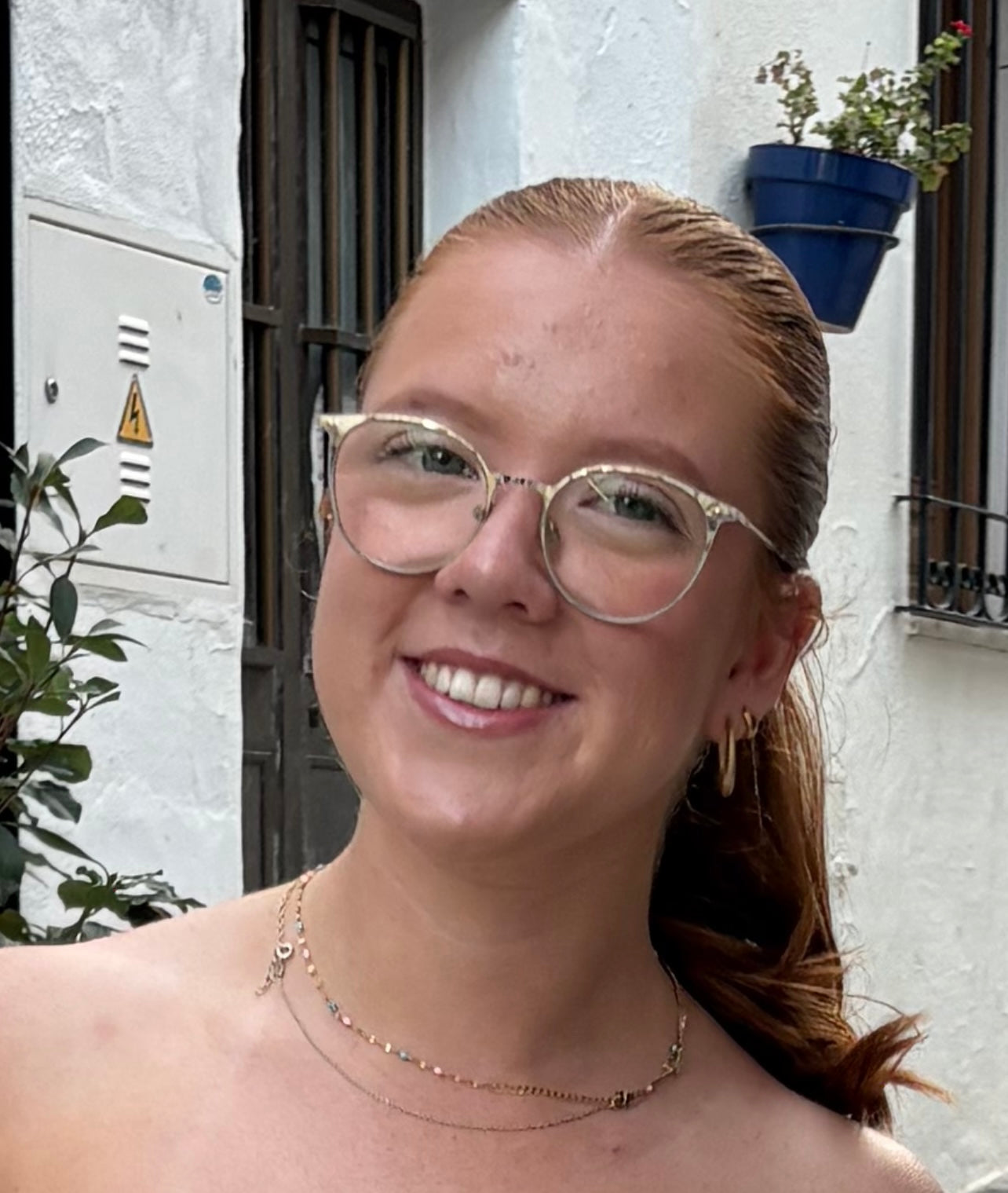 Paige, a woman with ginger hair wearing glasses with a white frame and gold jewellery standing in front of a white building. 