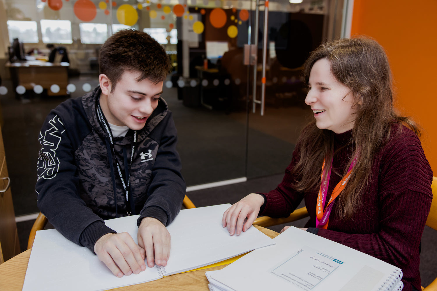 Two individuals seated at a table reading Brallie documents together in a modern office setting with glass walls and colorful decor.