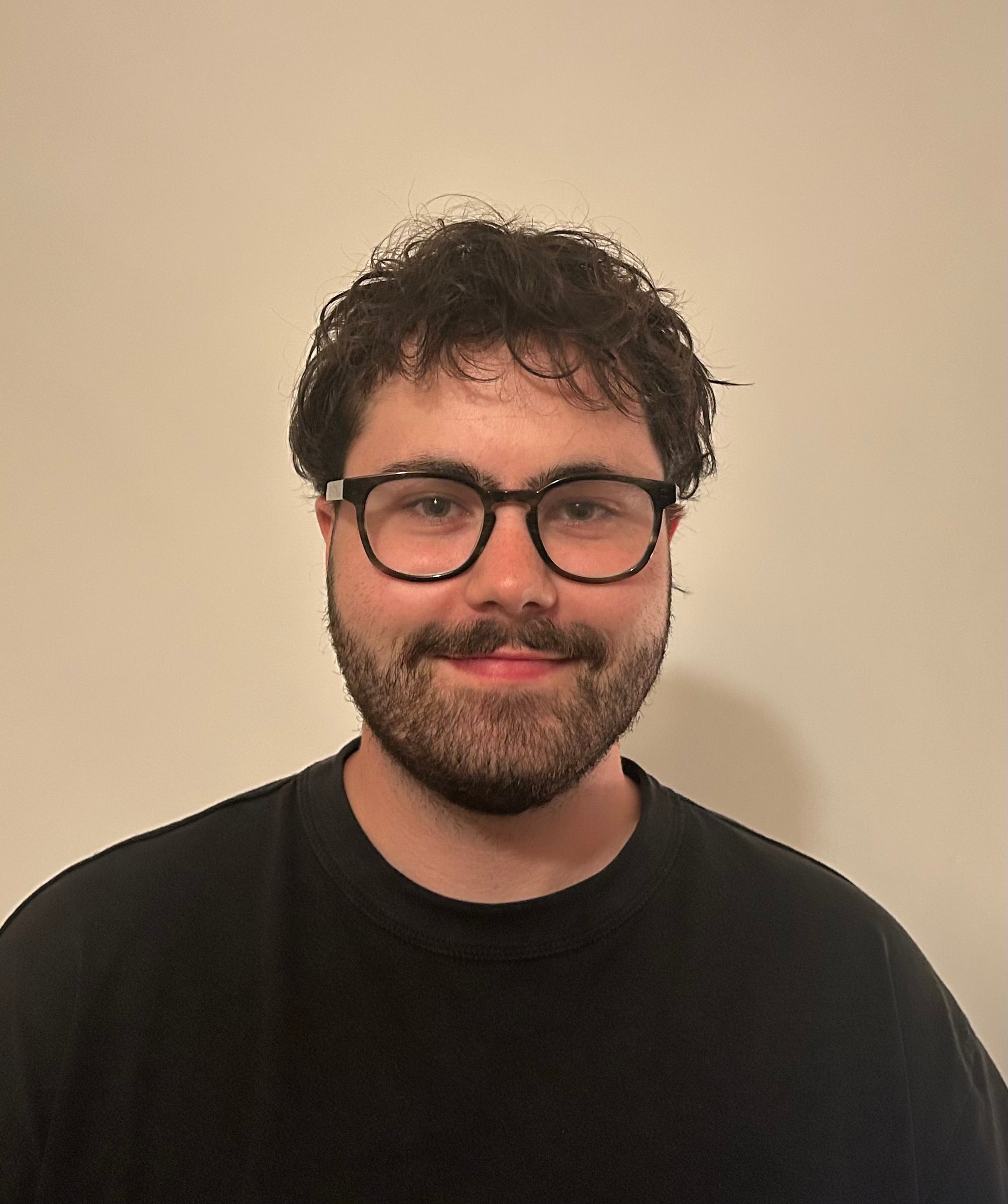 Seamus, a man with a short beard and mustache smiling against a cream wall. He's got short brown wavy hair and is wearing a black top and black circular glasses. 