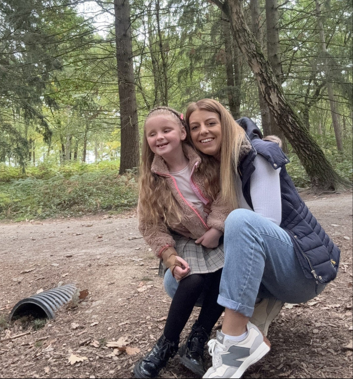 A woman crouching and hugging a young girl in a forest clearing surrounded by tall trees and greenery.