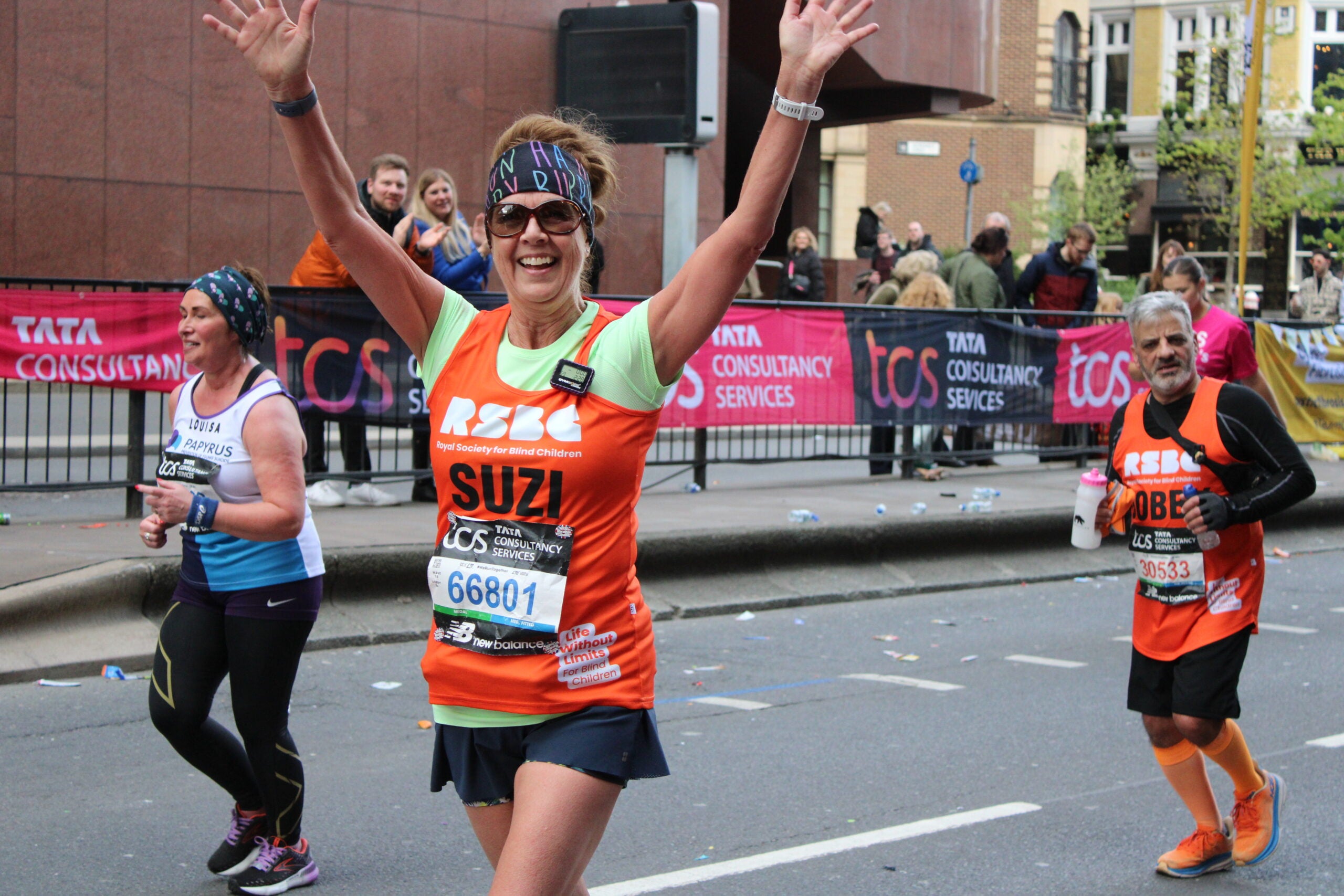 Suzi, a runner in a vibrant headband and orange RSBC running vest, has her hands in the air waving with a big smile as she is nearly at the finish line