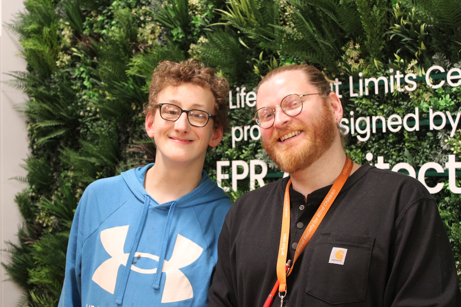Two people standing and smiling at the camera in front of a green lush wall made up of fake leaves. 