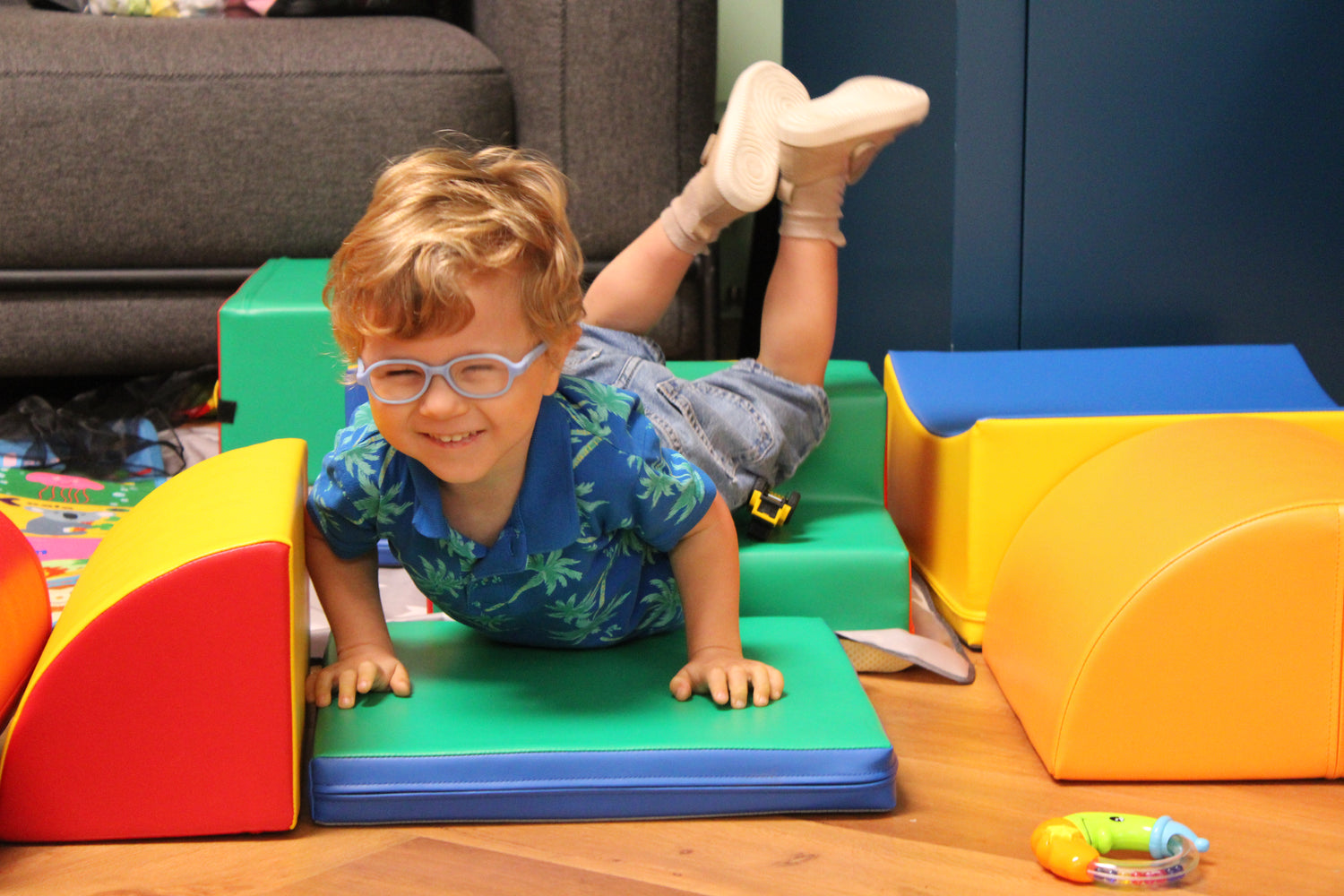 A young child in a blue floral shirt and denim shorts playing on colorful foam mats and cushions indoors.