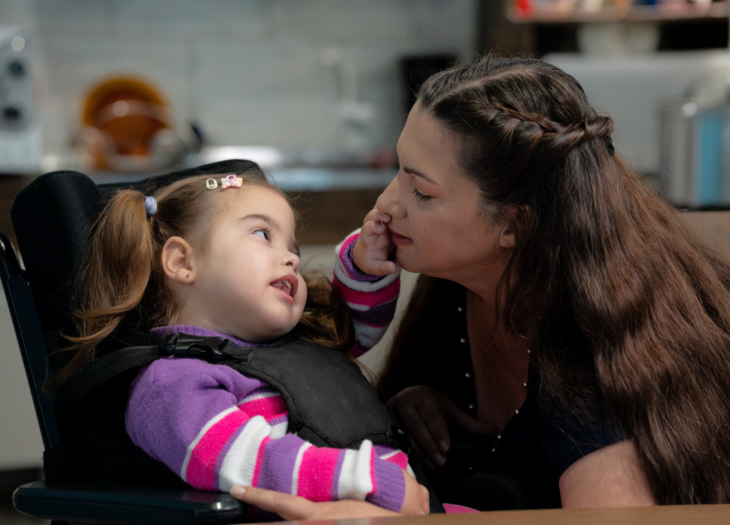 Mother sitting with her child who's in a high chair in a kitchen setting. The child is wearing a pink, white and purple stripped jumper and mother a black v-neck top with silver beads around the neckline.