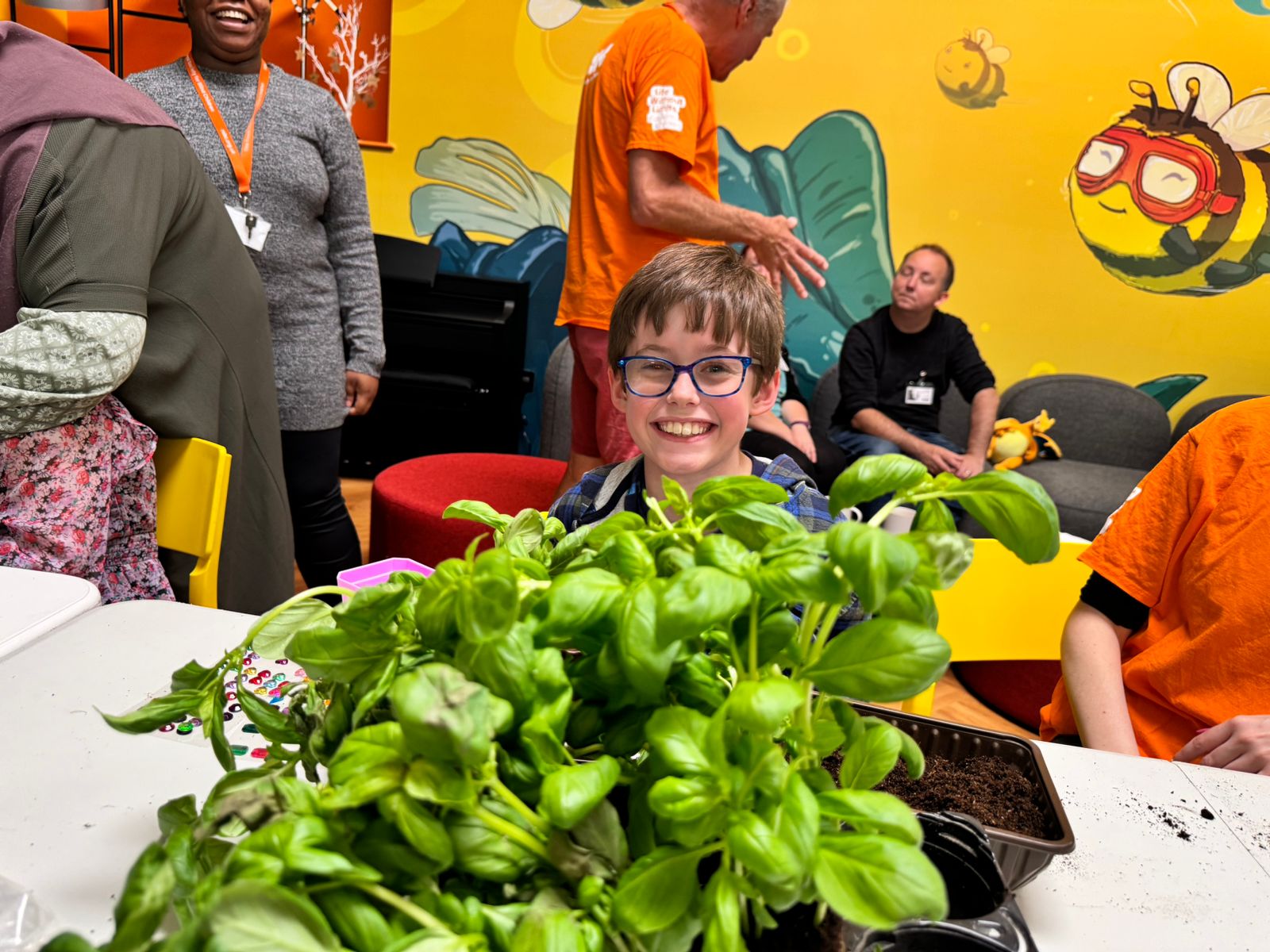A smiling young boy with glasses is behind a vibrant green plant in a busy gardening activity event