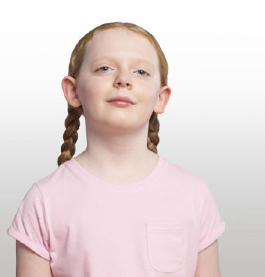A blind girl stands against a white backdrop. She has red hair which is platted and is wearing a pink t-shirt