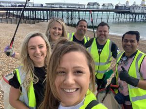 A group take a selfie on a beach with a pier in the background. They are wearing yellow hi vis jackets.