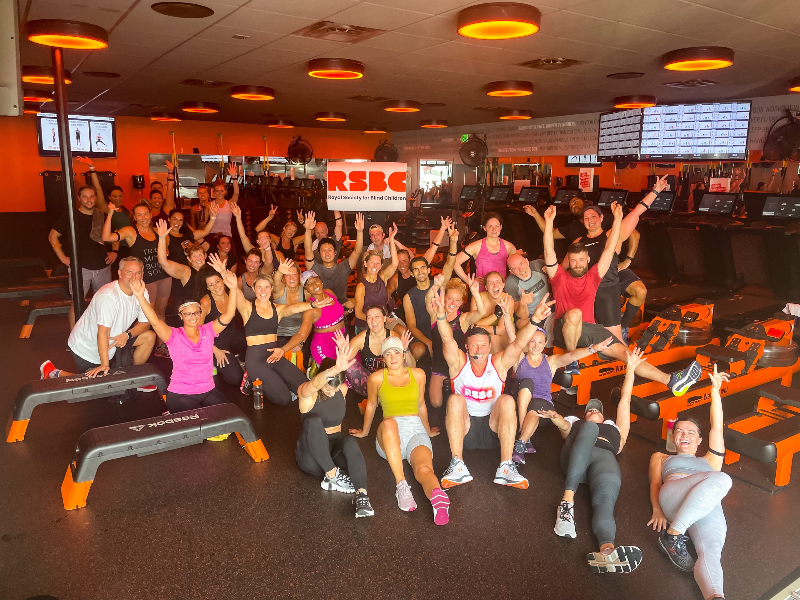 A large group of people in workout gear are gathered in a fitness studio, raising their arms and holding up a sign with the RSBC logo on.