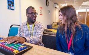 A young man is being taught how to use an accessible keyboard by his teacher. They are sitting next to each other at a desk and smiling.