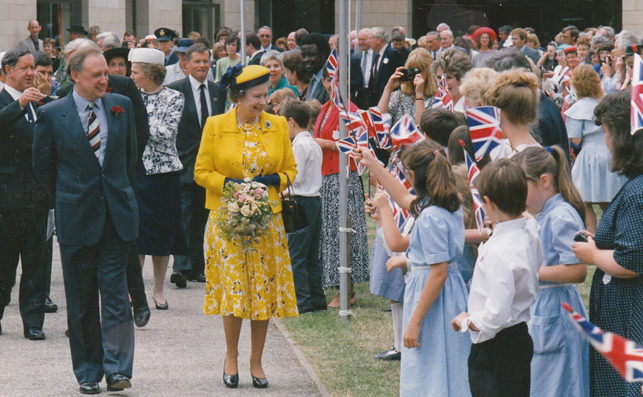 Queen, dressed in yellow, greets a line of children holding Union Jack flags at the opening of Dorton College.