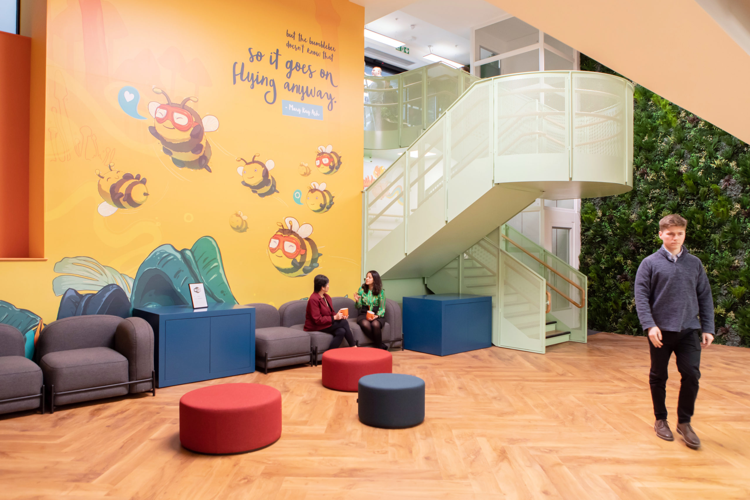 Two people sit on a sofa in a large atrium. There are seating stools in front of them, a colourful orange graphic on the wall behind them and a green staircase leading down from the entrance above