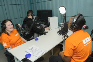 Two women are sitting in a soundproofed room wearing headphones and recording a podcast. There is a man behind them mixing using equipment.