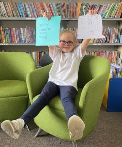 Abi holds up two pieces of paper. One is a letter to the Queen and the other is a picture of her in Buckingham Palace. 