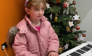 A girl playing piano next to a Christmas tree